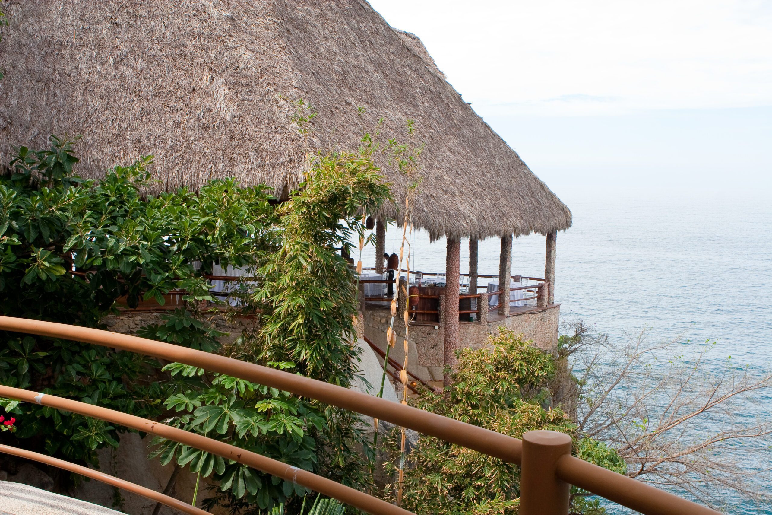A thatched-roof restaurant built on a cliff overlooking the ocean. There are tables set up on the outdoor patio, with a wooden walkway leading down to the water.