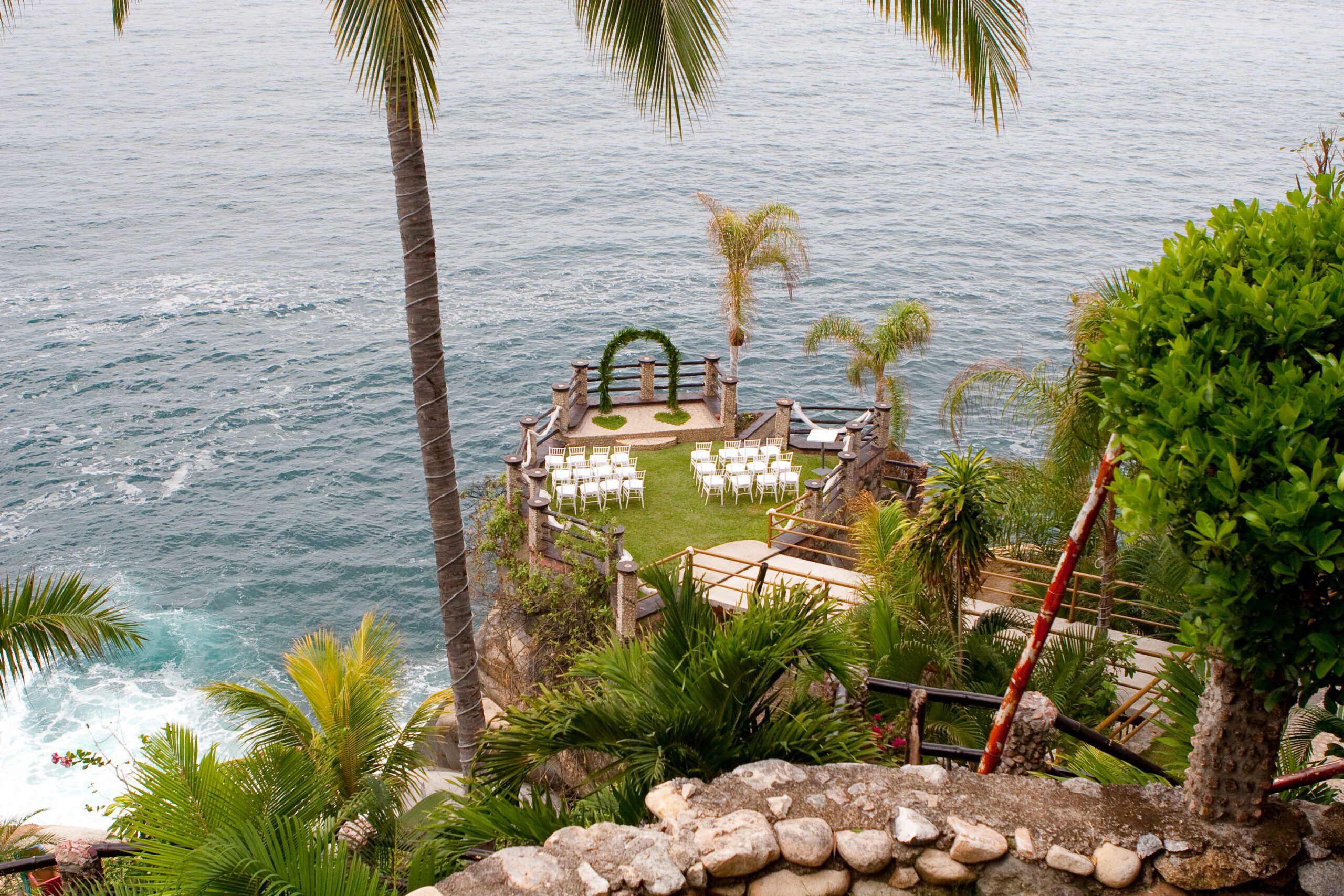 A picturesque cliffside wedding setup overlooking the ocean. The small, grassy platform features white chairs neatly arranged in rows and a circular green arch as a backdrop for the ceremony. Palm trees surround the area, adding a tropical atmosphere, while the ocean waves crash against the rocks below. The scene is framed by lush greenery and a stone wall in the foreground.