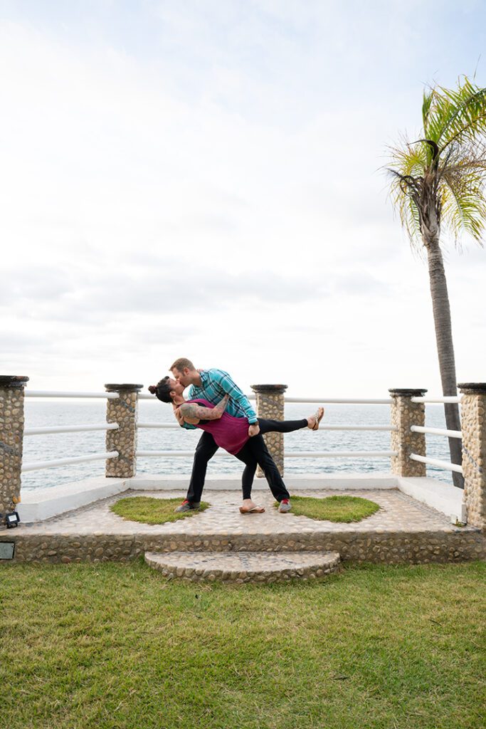 This is a photo taken in 2024 of Anna myself back at Le Kliff. If you walk down from the restaurant to the very bottom, they have a spot designated for weddings. At the very edge of the spot, they have two pieces of grass in the shape of a heart. In the background is the ocean with a palm tree on the left side. I am dipping Anna and I'm giving her a kiss at the same place we were married fifteen years ago.