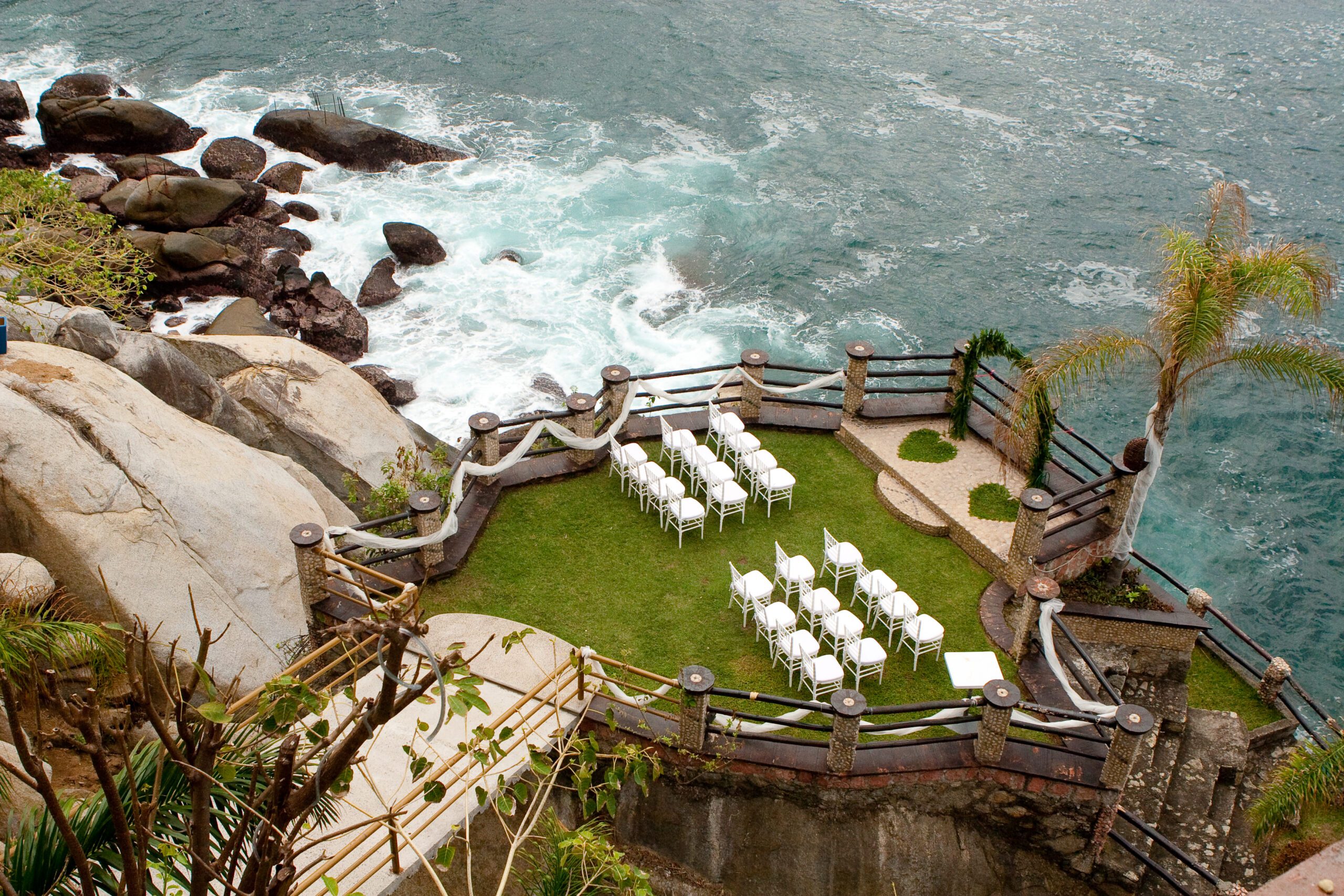A scenic outdoor wedding setup on a grassy terrace overlooking the ocean. Rows of white chairs are arranged in two sections facing an arch adorned with greenery. At the front of the chairs is two grass hearts for the bride and the groom. The terrace is surrounded by a stone and wooden railing, with waves crashing against rocky cliffs below. A palm tree stands to the right, adding to the tropical atmosphere.