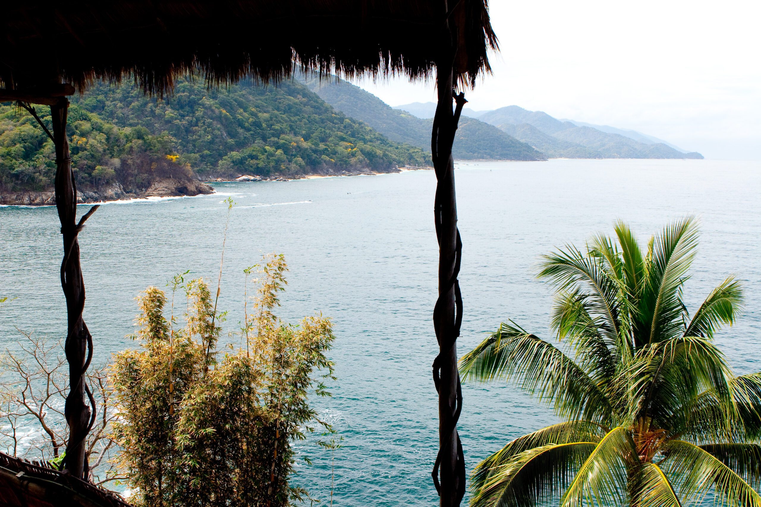 This is a view from the restaurant out on to the ocean. We can see the thatch roof at the top left with a palm tree at the bottom right. In the center is a view of the ocean and mountains on the left.