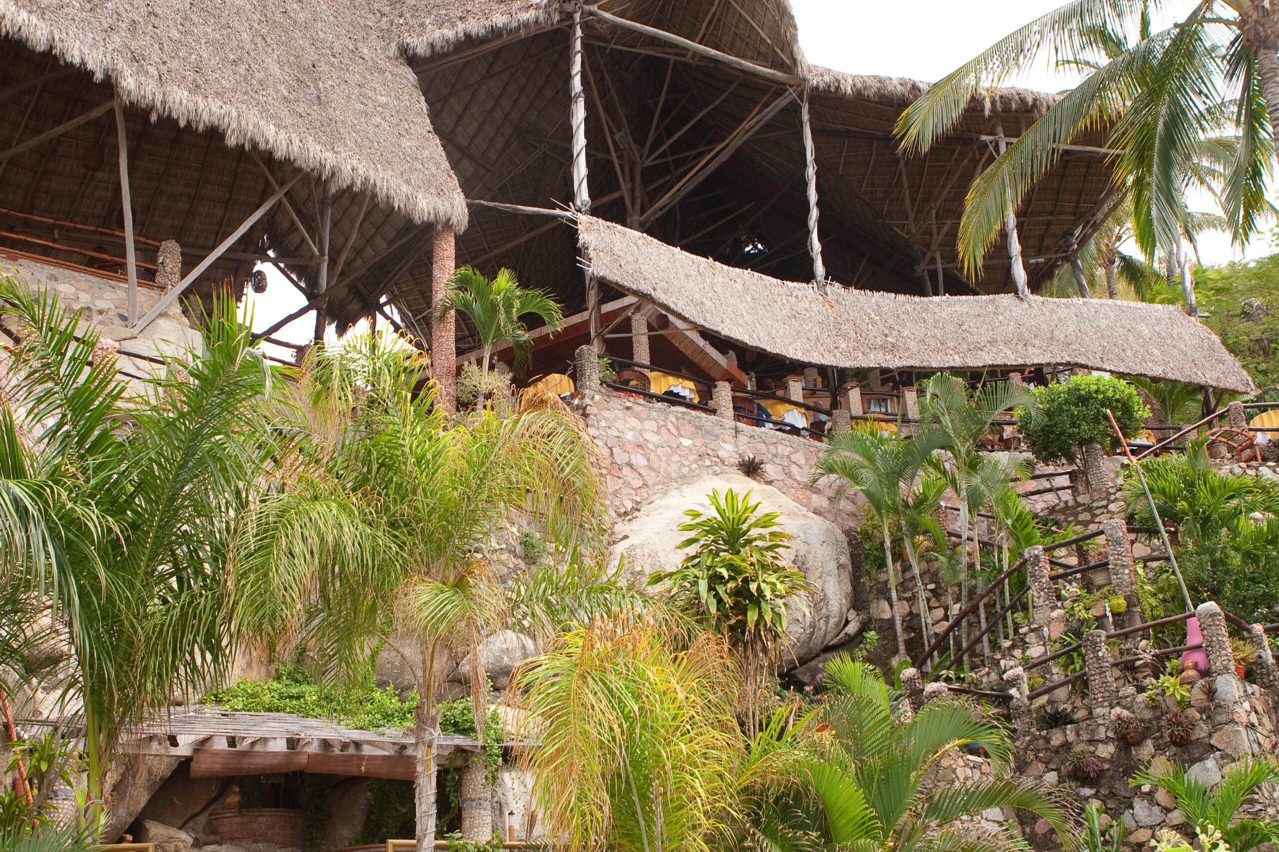 This is a picture looking from the wedding venue up to the restaurant. A large, multi-level open-air restaurant with a thatched roof built on a rocky hillside, surrounded by lush green palm trees and tropical plants. Stone pathways and wooden railings lead up the slope to the dining area, which overlooks the natural landscape. The structure features rustic wood beams, stonework, and natural materials that blend into the environment.