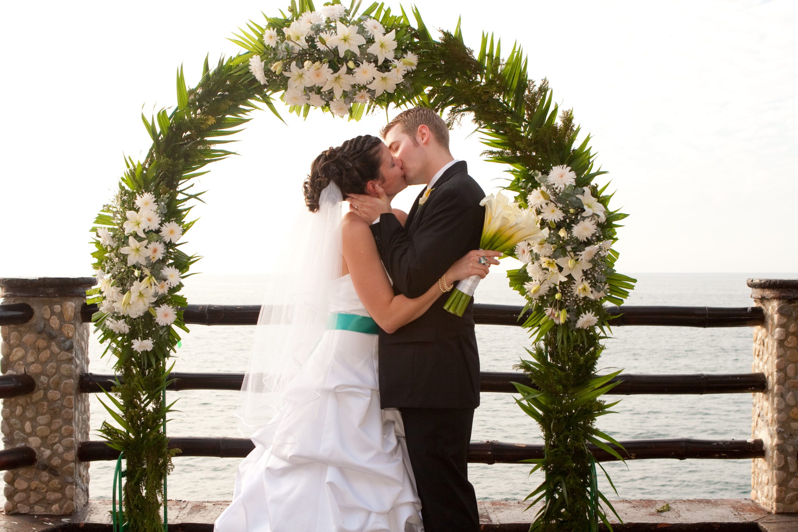 Photo of us kissing at the wedding venue. Anna is holding her bouquet in her right hand. Behind them is an ocean view.
