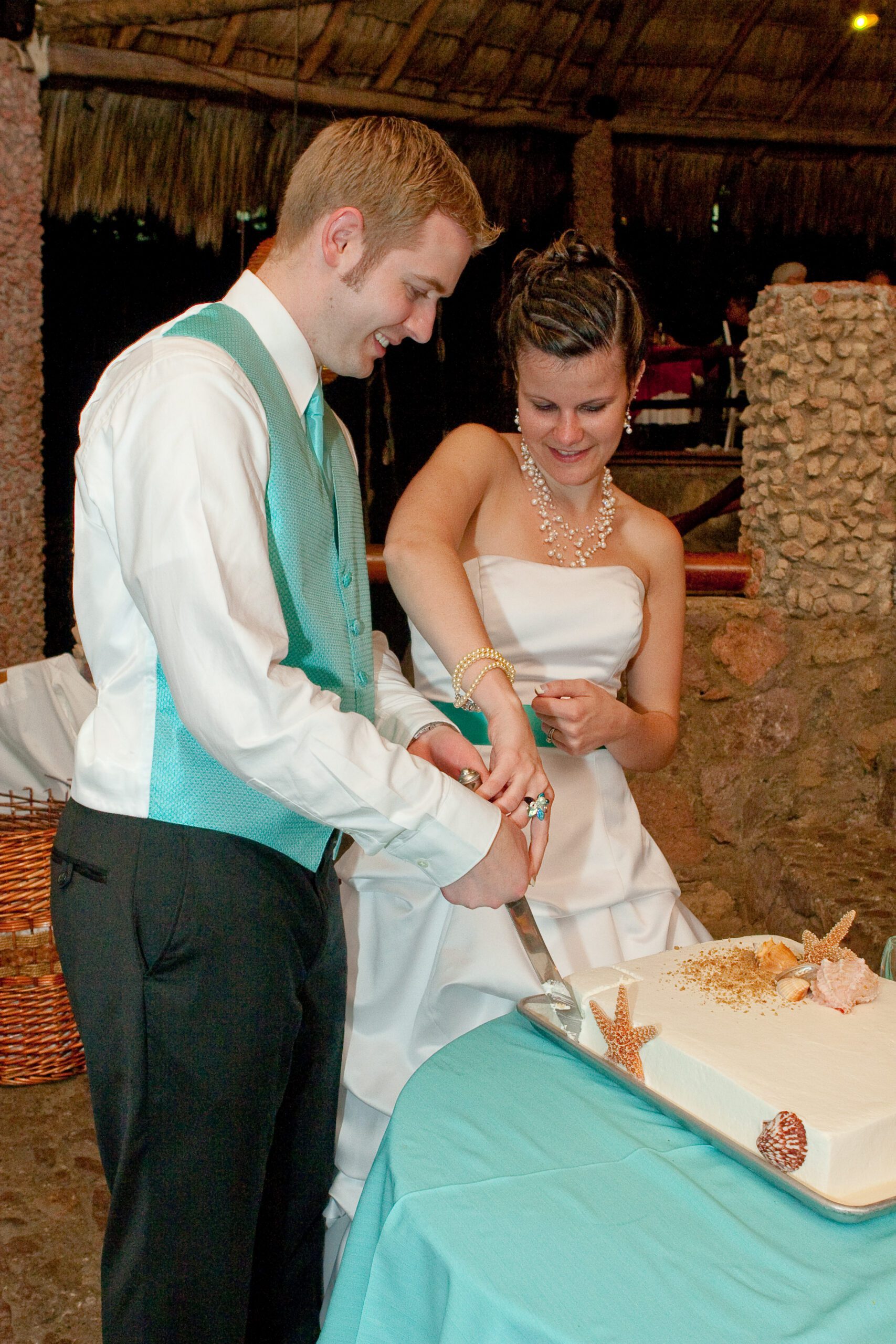 This is a picture of Adam and Anna cutting the tres leches cake. This was one of those moments where we had no idea what we should do even though we had seen other people cut wedding cakes before. Adam is in the foreground and Anna is in the background they're making their second cut to make a square out of this .