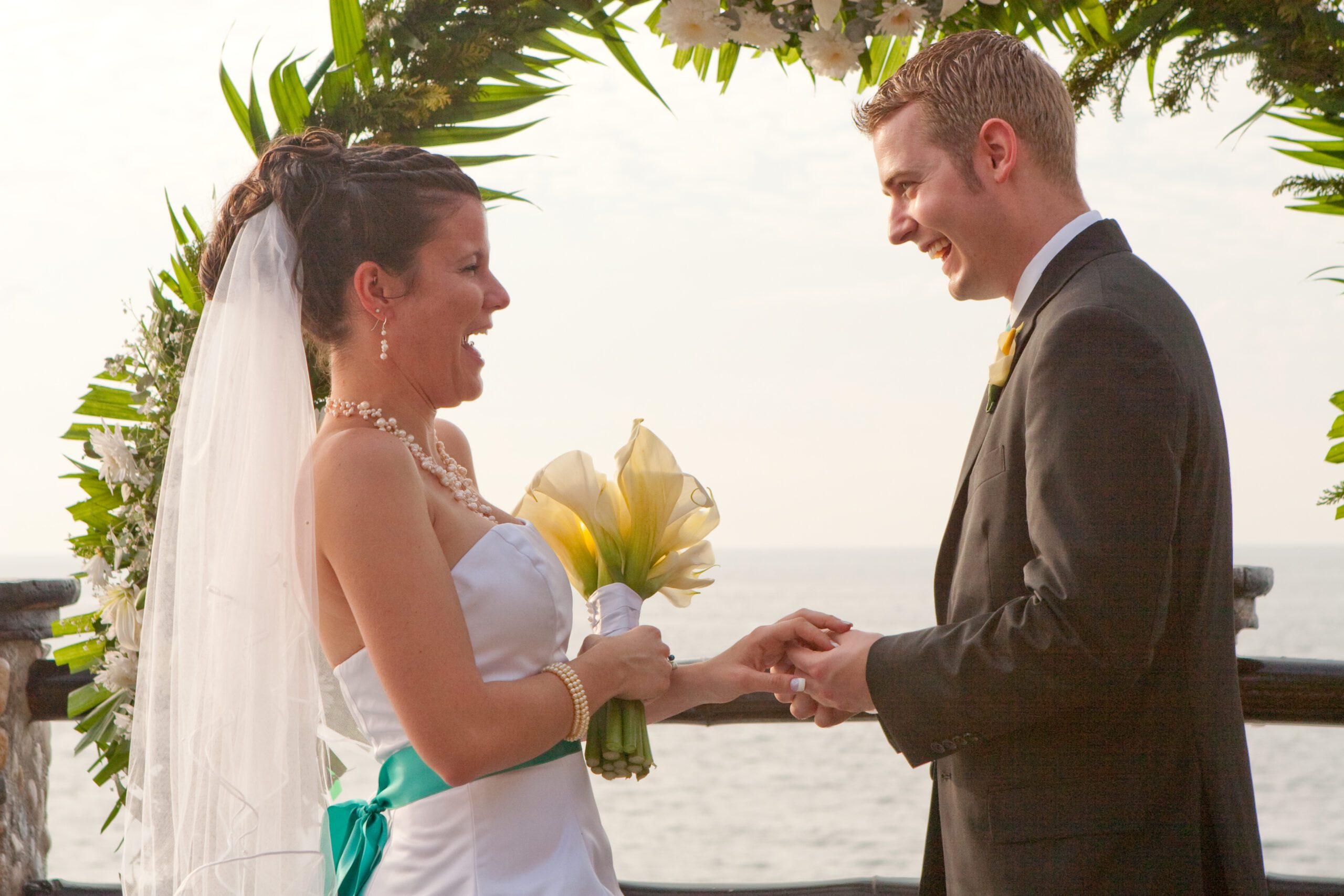 This is a photo of Adam and Anna laughing at the wedding venue right after Anna tried to put on Adam's ring and his fingers were swollen. There is an ocean in the background.