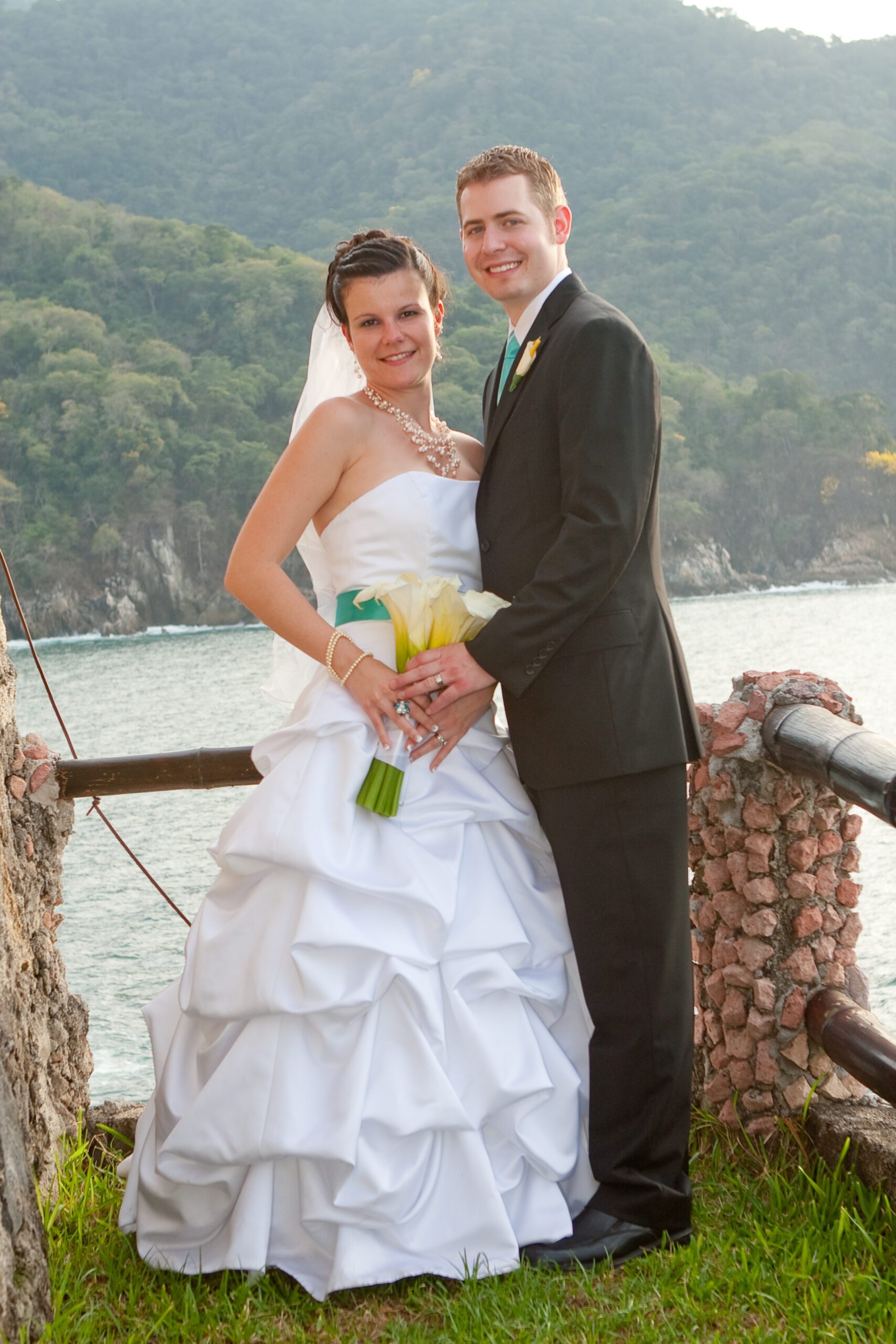 Vertical photo of Adam and Anna with the ocean and mountains in the background. Their hands are on Anna's right hip and she is holding the bouquet.