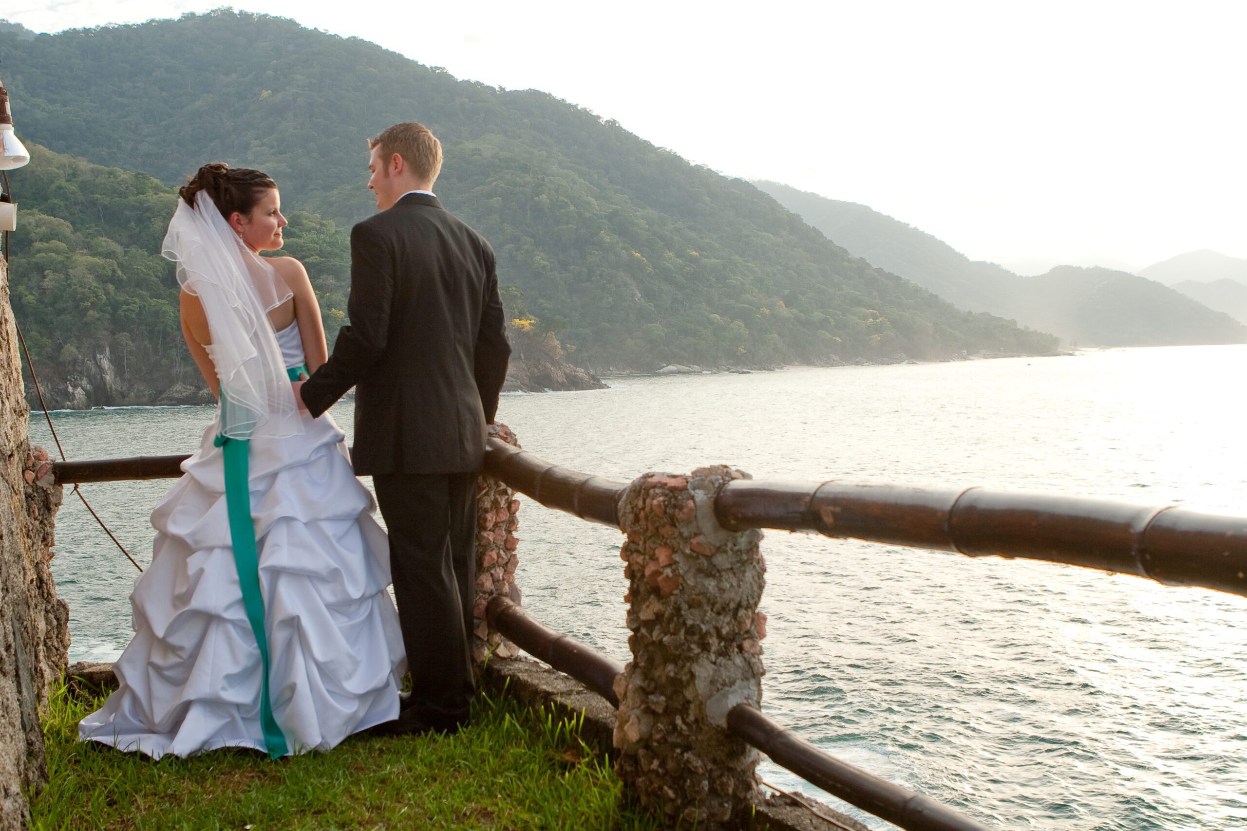 Anna's and Adam's back is to the camera. Adam's hand is on the small of Anna's back as they are looking at each other with the ocean and mountains in the background.