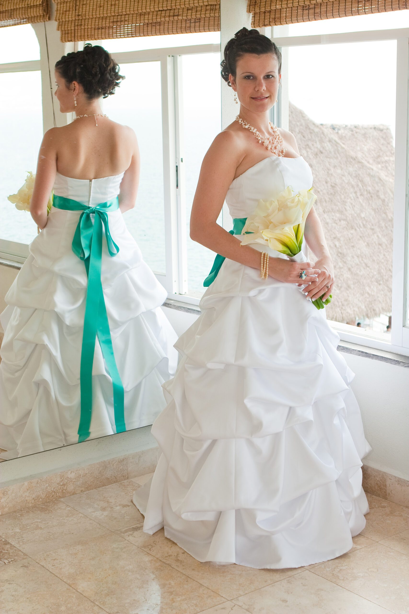 This is a vertical photo of Anna looking at the camera. Behind her is a mirror so you can see both the front and the back of her wedding dress. She is wearing a white cupcake dress that is strapless with a turquoise sash around her waist.