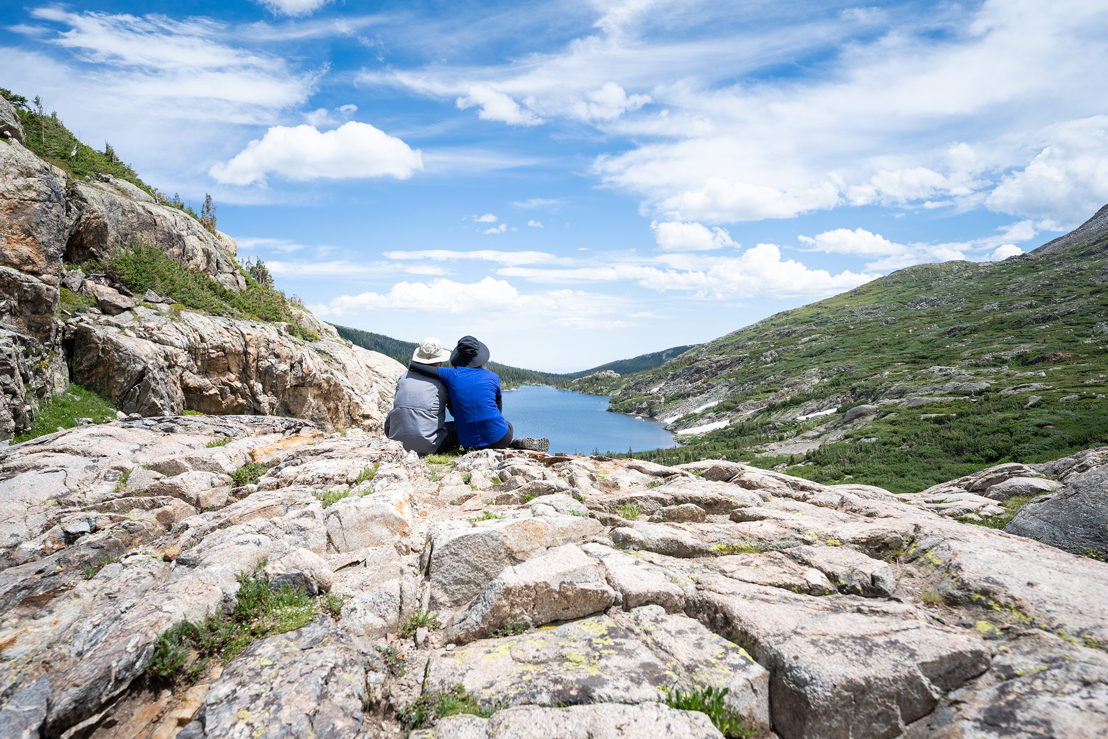 This is a photo of Anna and Adam sitting off in the distance at the top of one of their hikes. The camera was set up on a tripod and we can see them about middle frame. And it has her left arm wrapped around Adam a lake on a fairly cloudy day. On the left and right side, we can see the valley of the Rocky Mountains.